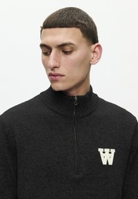 Pulover negru tricotat cu fermoar, având un guler ribbat, cu un logo brodat "W" de culoare bej pe partea stângă. Textură netedă, design casual.