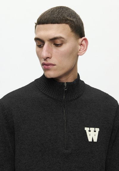 Sweater noir en tricot à fermeture éclair avec un col côtelé, arborant un logo "W" beige brodé sur le côté gauche. Texture lisse, design décontracté.