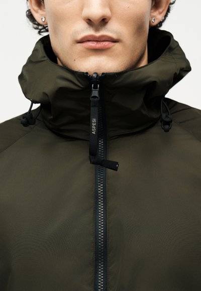Gros plan sur une personne portant une veste à capuche vert foncé avec une tirette de fermeture éclair noire portant l'inscription "ASPESI" et des cordons de serrage sur la capuche.