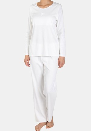 Person trägt ein schlichtes weißes Langarmshirt und passende weiße Hose, steht barfuß vor neutralem Hintergrund.