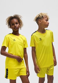 Żółte sportowe koszulki i spodenki z lekkiego materiału. Czarne akcenty na ramionach i spodenkach z wzorem w jodełkę.