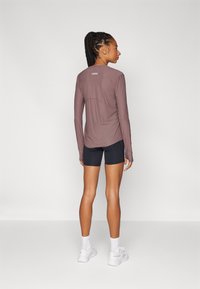 Camiseta deportiva de manga larga en color malva con dobladillo redondeado, combinada con pantalones cortos negros. Logo blanco en la parte de atrás, tejido suave, estilo ajustado.