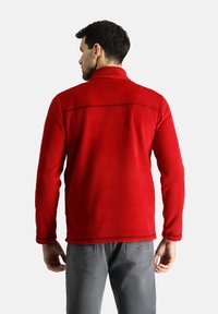 Chaqueta de forro polar roja con un cuello alto, presenta una costura trasera recta y un pespunte en contraste. Combinada con jeans grises.