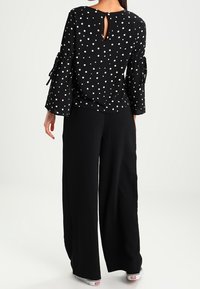 Black blouse with white polka dots, 3/4 bell sleeves with ties, keyhole back detail. Paired with loose-fitting black pants and white sneakers.