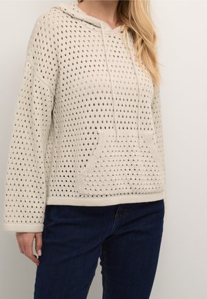 Femme portant un sweat à capuche en tricot de couleur crème avec un motif perforé et une poche kangourou à l'avant, associé à un jean bleu foncé.