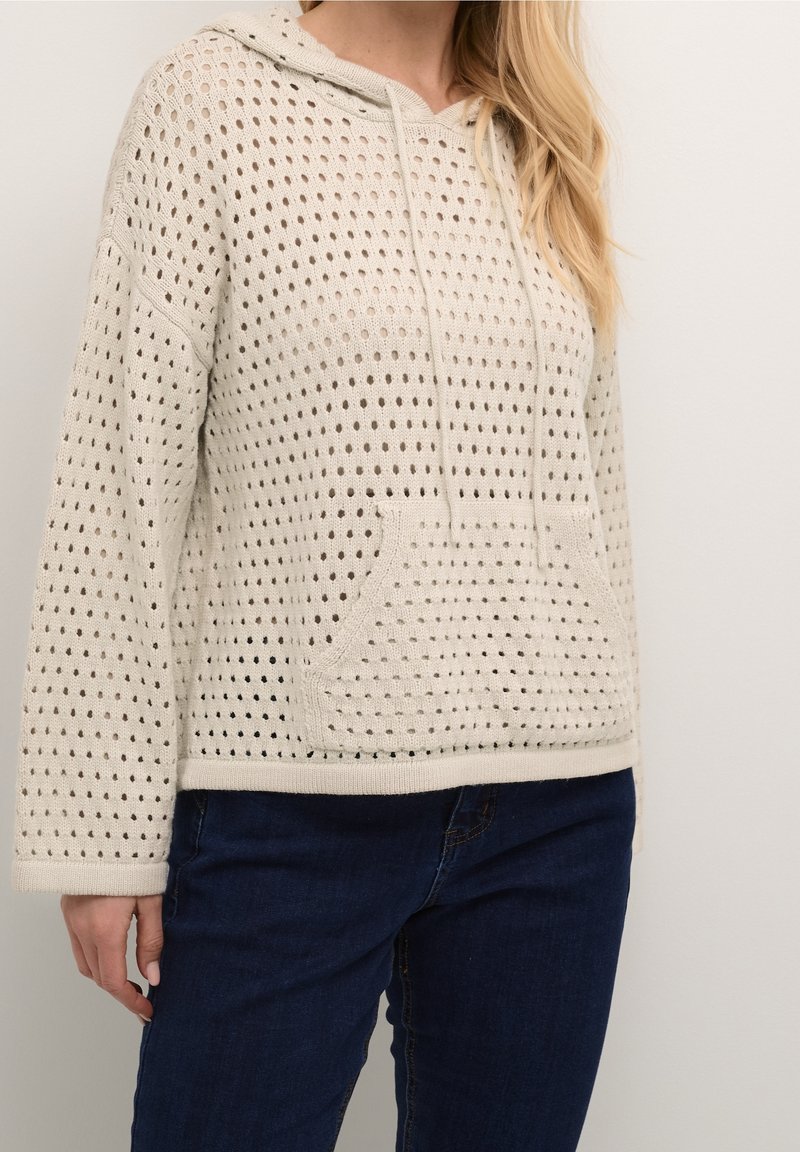 Femme portant un sweat à capuche en tricot de couleur crème avec un motif perforé et une poche kangourou à l'avant, associé à un jean bleu foncé.