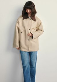 Kobieta z ciemnymi włosami do ramion, ubrana w beżową kurtkę zapinaną na guziki z ciemnogranatowym kołnierzem i niebieskimi dżinsami, uśmiechająca się i patrząca w dół.