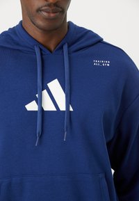 Modrá mikina s kapucňou a bielym logom Adidas. Obsahuje šnúrky a text "TRAINING ALL_GYM" na ramene. Mäkko tkanina, ležérny dizajn.