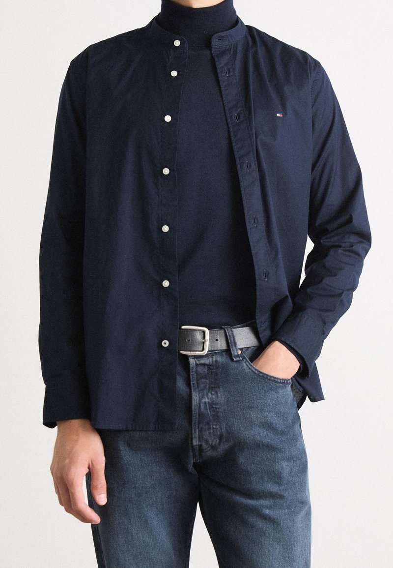 Homme portant une chemise bleu marine boutonnée sur un col roulé bleu marine, un jean bleu avec une ceinture noire, une main dans la poche, debout devant un fond blanc.