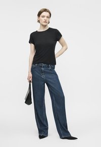 Femeie purtând un tricou negru și blugi largi albaștri, ținând o geantă neagră într-o mână, stând pe un fundal deschis și simplu.