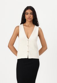 Kremowy, bezrękawnik dzianinowy z głębokim dekoltem w kształcie litery V, z pięcioma guzikami z przodu i fakturowanym wykończeniem. Połączony z czarną spódnicą.