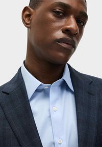 Blauw overhemd onder een donker geblokte blazer. Het overhemd heeft een spread collar en een knoopsluiting met lichtgekleurde knopen.