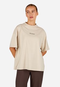 Bežs pārmērīgs t-krekls ar īsām piedurknēm un apaļu kakla izgriezumu, priekšpusē ar diskretu melnu "PEGADOR" logotipu. Gluda kokvilnas tekstūra.