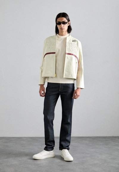 Cremefarvet jakke med præget logo, bordeaux accenter, lynlås-lommer; beige top; mørke jeans; hvide sneakers. Glat tekstur, cropped design.