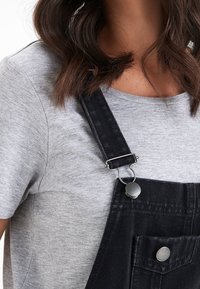 T-shirt gris associé à des salopettes en denim noires. La salopette présente des clips en métal argenté et des accents de boutons sur les poches.