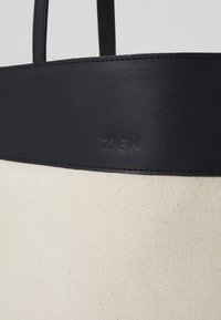 Sac en toile avec un corps crème et un dessus en cuir noir. Présente un logo "ZIGN" en relief et des coutures ton sur ton. Design minimaliste.