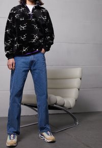 Personne portant un polaire à motifs noir et blanc, un jean bleu et des baskets beiges se tient à côté d'une chaise blanche moderne et rembourrée sur un sol gris.