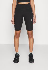 Kvinna som bär svarta högmidjade cykelshorts från Adidas och svarta sneakers med ljusgröna sockor, stående mot en enfärgad vit bakgrund.