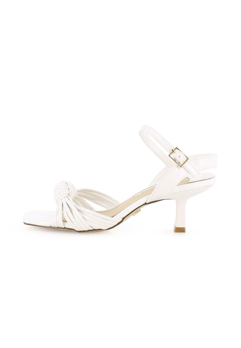 Sandalia blanca de tacón con diseño frontal anudado, tiras estrechas, parte trasera abierta y tacón de 7.5 cm. Textura suave con acabado brillante y detalles en dorado.