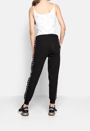 Black joggers with a tapered fit, elastic waistband, and side stripes featuring a white graphic pattern. Paired with black shoes.