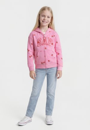 Ragazza sorridente con felpa rosa GAP con stampe di fragole, jeans azzurri e sneaker bianche, in piedi su sfondo bianco.