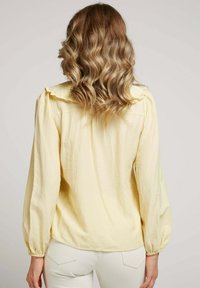 Blouse jaune avec un col volanté, des manches ballons et un tissu texturé. Portée avec un pantalon clair. La vue de dos montre des cheveux tombant sur les épaules.