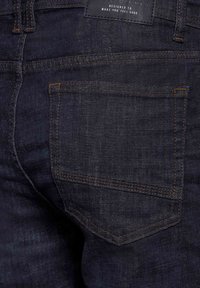 Tmavé džínové kalhoty s zadní kapsou s oranžovým švem a texturovaným povrchem. Štítek na pase s vytlačeným textem.