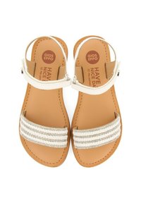 Sandalias planas con suelas de cuero marrón. Las tiras blancas cuentan con tres filas de acentos metálicos y una correa en el tobillo con una hebilla plateada.