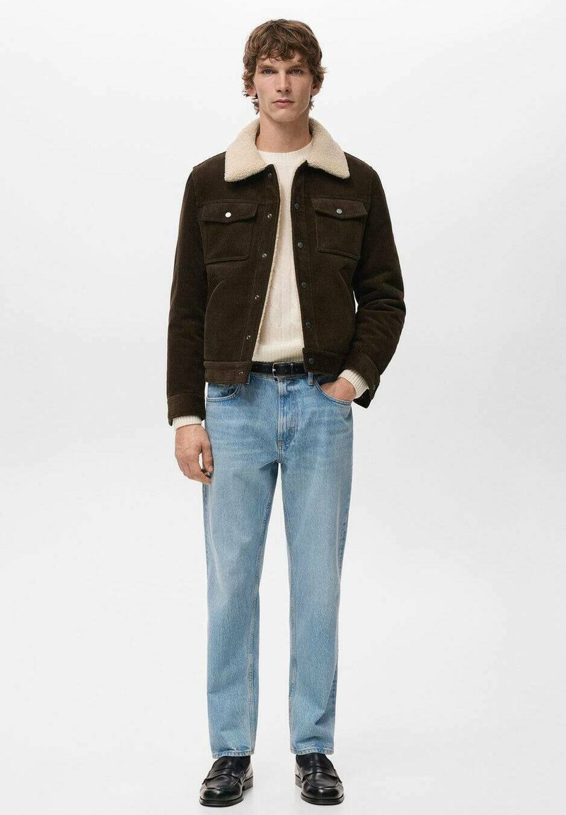 Giacca in velluto a coste marrone con colletto crema, due tasche sul petto, abbinata a jeans azzurri e mocassini neri. Design semplice e classico.