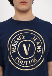 Tumši zila kokvilnas T-kreklu ar zelta apaļu logotipu, uz kura ir uzraksts "VERSACE JEANS COUTURE" un izcelts "V" centrā. Apaļš kakls.