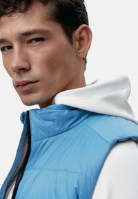 Gilet matelassé bleu clair avec un col haut, une fermeture éclair et une texture matelassée. Porté sur un sweat à capuche blanc avec une capuche visible.