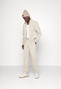 Completo tuta beige con giacca zip-up e pantaloni con coulisse, con dettagli del logo verde sulle maniche e sulle gambe, completato da un berretto beige.