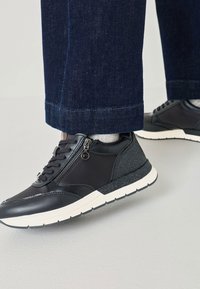 Čierne tenisky z hladkej kože a textilného materiálu, so zipsom na boku, bielou podrážkou a šedými detailmi, zobrazené s tmavými denimovými nohavicami.