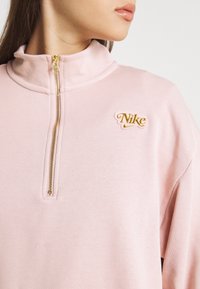 Sudadera de color rosa claro con cuello alto, con un cierre dorado y un logo dorado de "Nike". Textura suave, ajuste estándar y detalles acanalados.