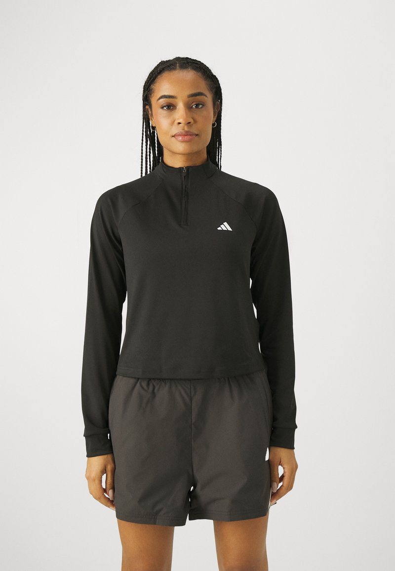 Maglia nera con zip e maniche lunghe, dotata di colletto alto, realizzata in tessuto morbido. Abbinata a pantaloni corti scuri, che mostrano una vestibilità rilassata. Dettaglio del logo bianco.