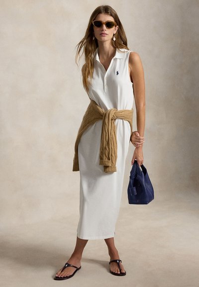 Femme portant une robe polo blanche sans manches avec un pull beige noué à la taille, tenant un sac à main bleu marine, portant des lunettes de soleil et des sandales noires.