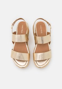 Sandales métalliques dorées avec deux larges brides et un design à talon arrière, dotées d'une semelle intérieure rembourrée couleur tan et d'une semelle de plateforme texturée.