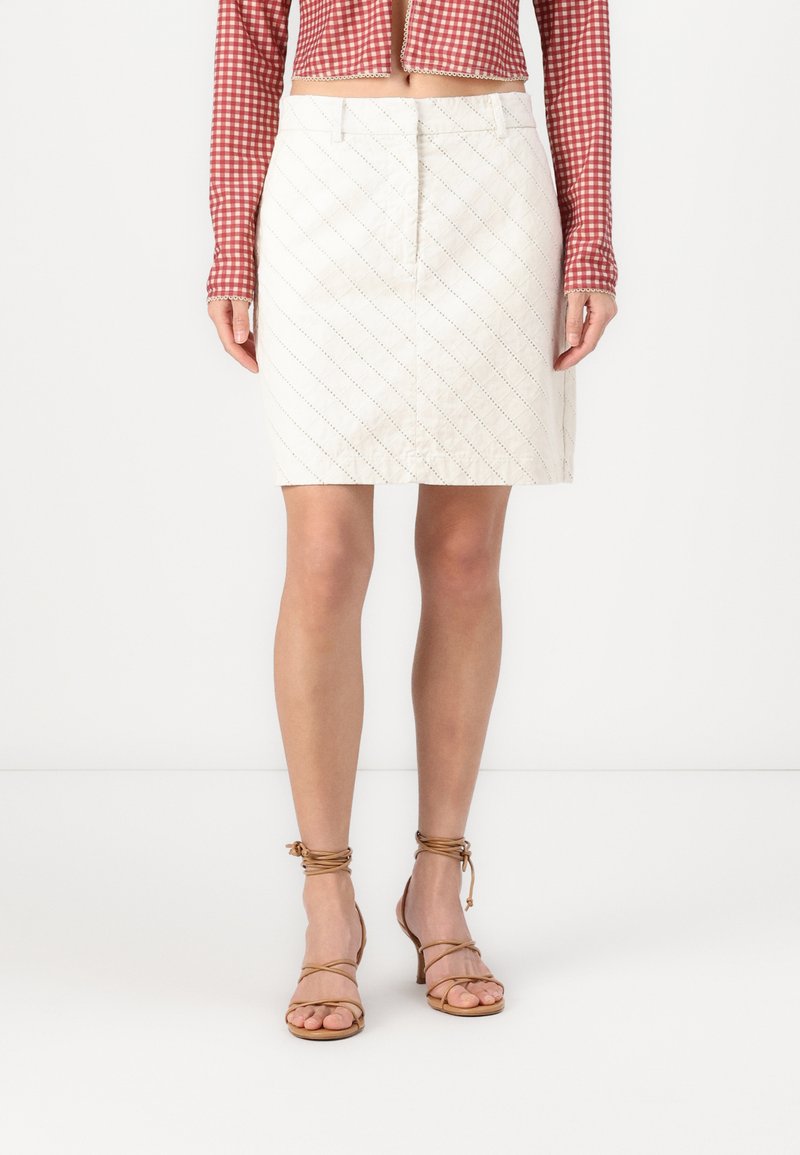 Λευκή φούστα με διακοσμητικό σχέδιο σε σχήμα διαμαντιού και διακριτική ραφή, συνδυασμένη με καφέ σανδάλια με λουράκια, αναδεικνύοντας τις ομαλές υφές και τη στενή γραμμή.