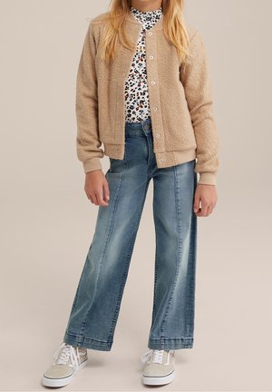 Beige fleece bomberjakke over en hvid skjorte med sorte og brune pletter, parret med brede blå jeans og hvide sneakers.