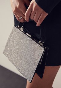 Rokassoma ar mirdzošu rhinestone pārklājumu, taisnstūra formu, melnas ādas akcentiem un divu rokturu dizainu ērtai nēsāšanai.