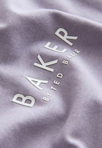 Liliowy materiał o teksturowanej powierzchni z wypukłymi, białymi literami: wyraźnie widoczne "BAKER" z mniejszym napisem "by TED BAKER" poniżej.