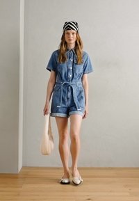 Denim jumpsuit med korte ermer og shorts, belte i midjen, lys blå farge, frontknapper, kombinert med et mønstret hodebånd og flate sko.