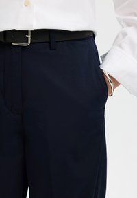 Άτομο που φοράει σκούρο μπλε παντελόνι, μαύρη ζώνη, λευκό πουκάμισο και ασημένιο βραχιόλι, με το χέρι στην τσέπη.