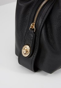 Musta teksturoitu nahkalaukku kulmassa, jossa on kultainen vetoketju ja pyöreä kultainen kiinnitysliitin hihnassa neutraalilla taustalla.