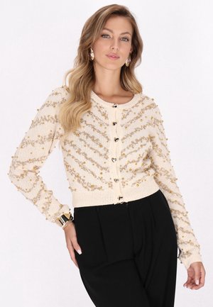 Perlenbesetzte cremefarbene Strickjacke mit einem Zopfstrickmuster und goldenen Akzenten. Verfügt über eine durchgehende Knopfleiste sowie gerippte Bündchen und Saum. Kombiniert mit schwarzen Hosen.