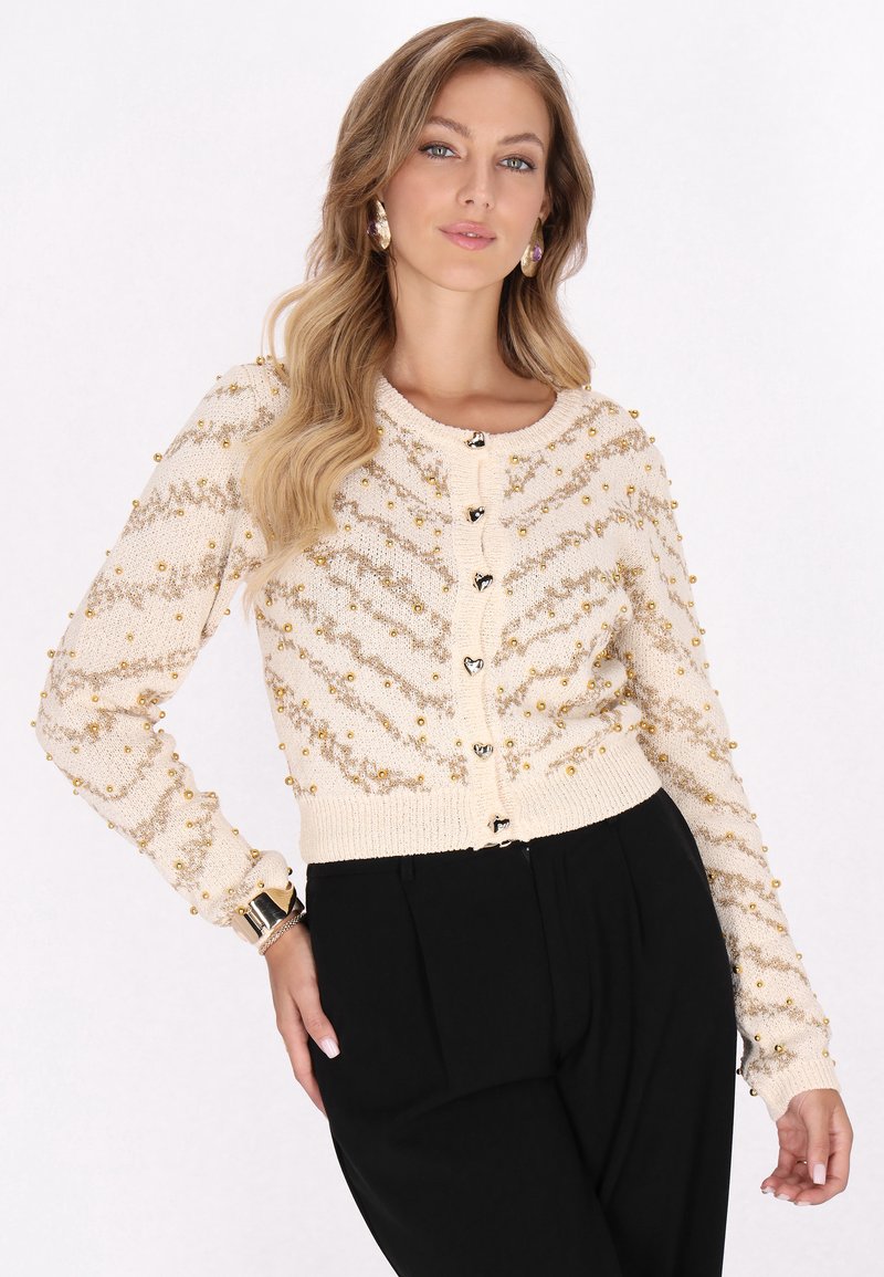 Cárdigan crema con cuentas, con un patrón de trenzas y acentos dorados. Presenta un cierre frontal con botones y un dobladillo y puños acanalados. Combinado con pantalones negros.