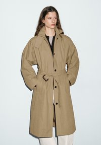 Manteau trench kaki avec taille ceinturée, grand col et deux poches latérales. Fabriqué en tissu respirant avec une coupe décontractée et une fermeture à boutons.
