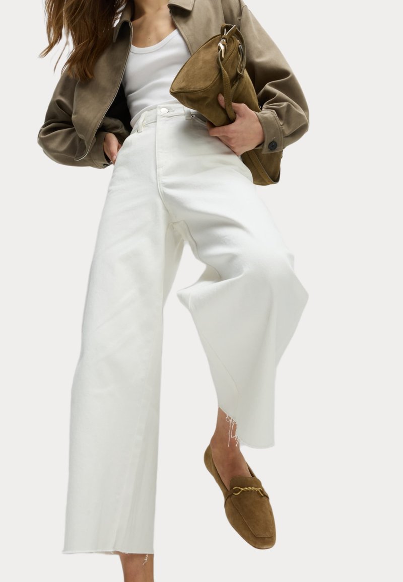 Jean large blanc, veste en suède beige et sac en suède marron, associés à des mocassins marron. L'ensemble présente des lignes épurées et des textures.