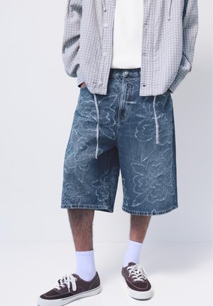 Mand iført batikmønstrede denimshorts, hvide sokker, mørke sneakers, hvid skjorte og grå ternet skjorte med knapper med hænderne i lommerne.