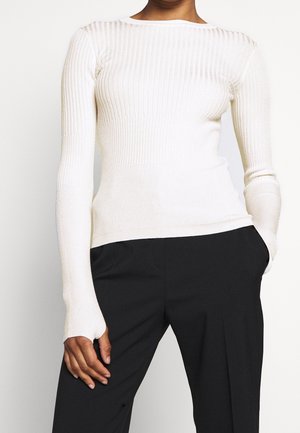 Femme portant un pull blanc côtelé ajusté à manches longues, associé à un pantalon noir ample, une main dans la poche, sur un fond uni.